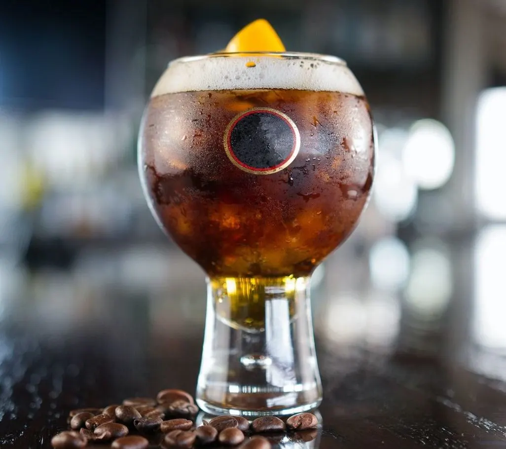 Ein kugelförmiges Glas mit dunklem Kaltgetränk, Eis, Schaum und Orangenscheibe. Es hat ein schwarzes Etikett mit rotem Rand. Dazu Kaffeebohnen auf dunkler Oberfläche.
