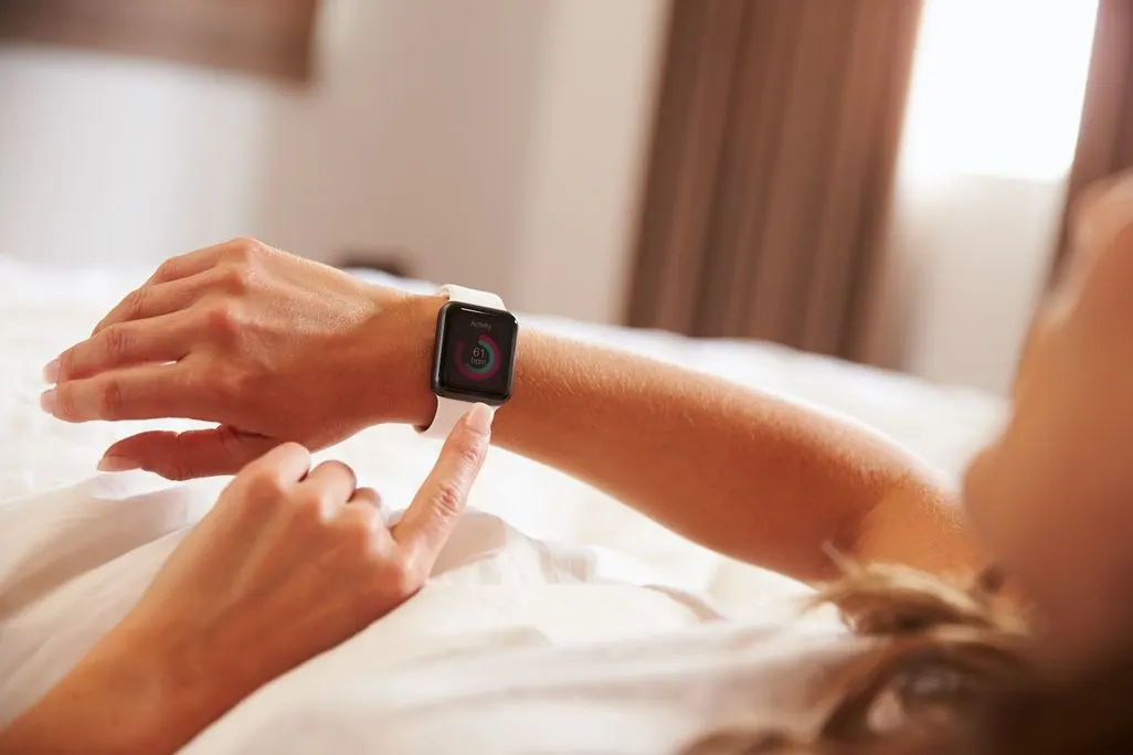 Person liegt im Bett und tippt auf eine Smartwatch mit weißem Armband.