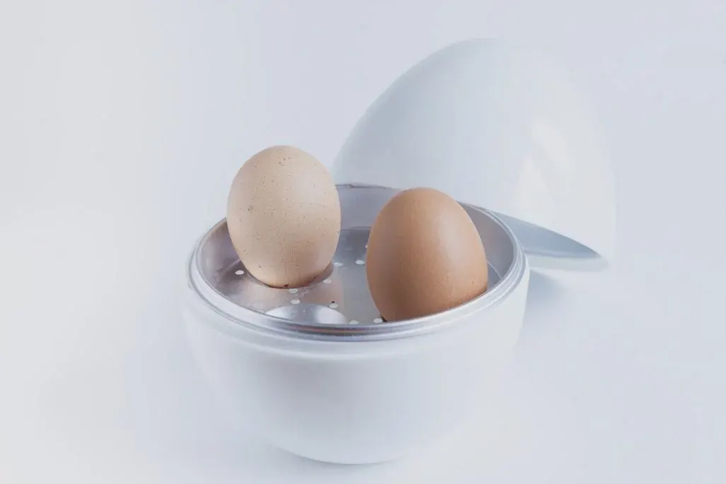 Zwei braune Eier stehen in einem kleinen weißen Eierkocher mit geöffnetem Deckel auf Metall-Einsatz, vor hellem, neutralem Hintergrund.