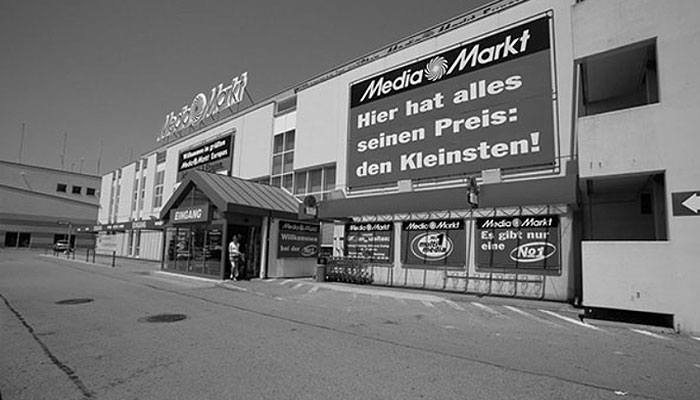 Eine Schwarzweißaufnahme eines MediaMarkt-Gebäudes mit einem großen Werbeplakat darüber. Der Eingangsbereich ist sichtbar.
