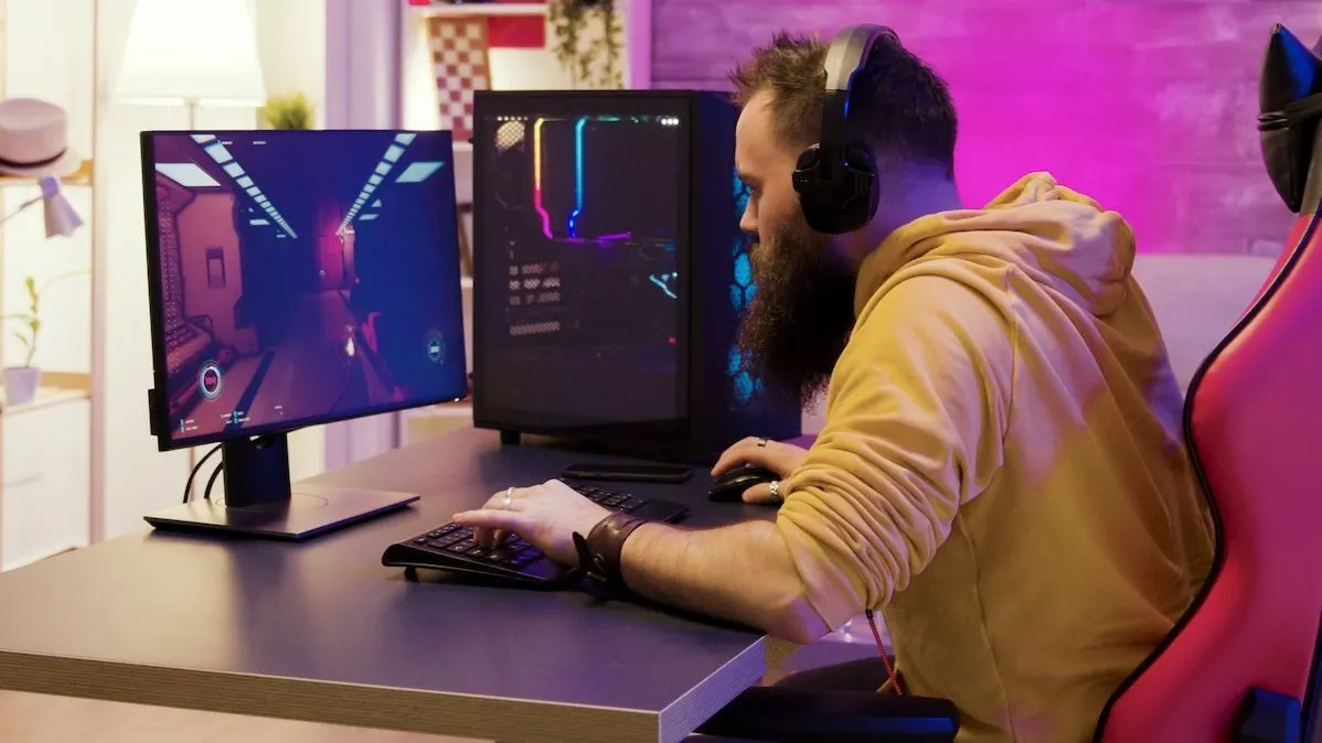 Ein Mann mit Kopfhörern sitzt an einem Gaming Schreibtisch und spielt ein Computerspiel auf einem PC und  Monitor