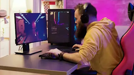 Ein Mann mit Kopfhörern sitzt an einem Gaming Schreibtisch und spielt ein Computerspiel auf einem PC und Monitor