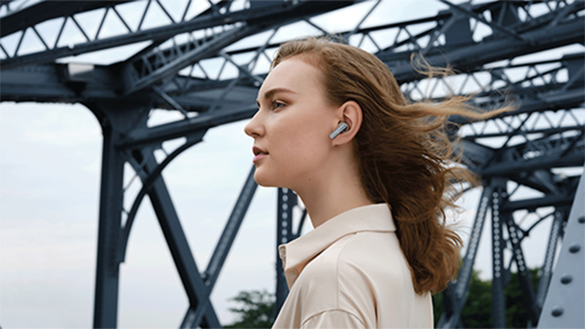 Eine Frau mit hellbeigem Hemd und Ohrhörern steht vor einer grauen Stahlbrücke.
