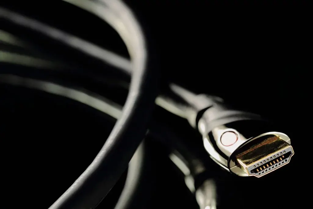 Ein HDMI-Kabel mit goldfarbenem Stecker, silbernem Gehäuse und weißem Punkt, vor dunklem, unscharfem Hintergrund.