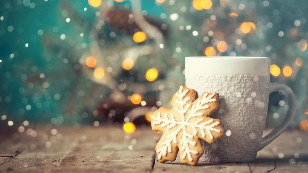 Weiße Tasse mit Schneeflocken-Keks vor winterlicher Lichterdekoration.