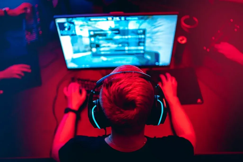 Person mit leuchtenden Kopfhörern spielt Videospiel an Monitor, Hände auf Tastatur/Maus. Rotes Umgebungslicht, weitere Hände sichtbar.