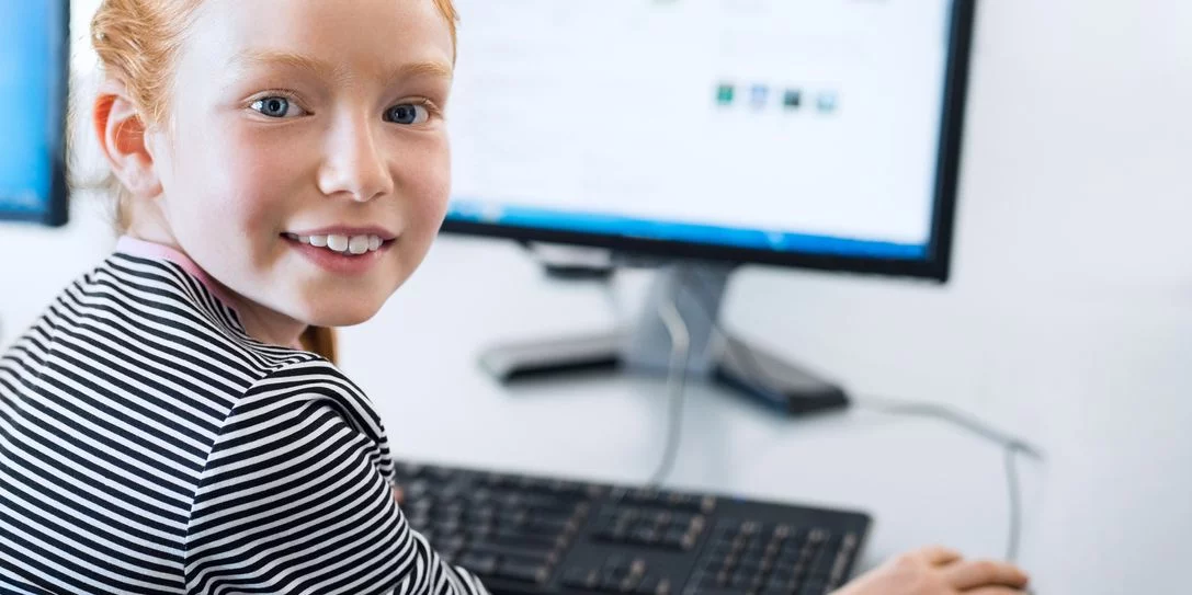 Lachendes Kind in gestreiftem Shirt am Computer mit Monitor und Tastatur.