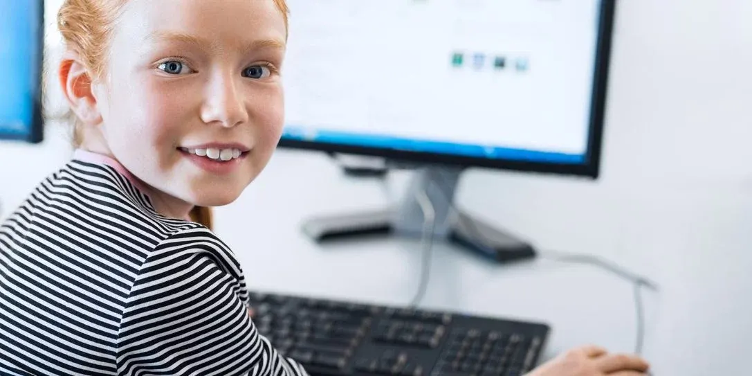 Lachendes Kind in gestreiftem Shirt am Computer mit Monitor und Tastatur.