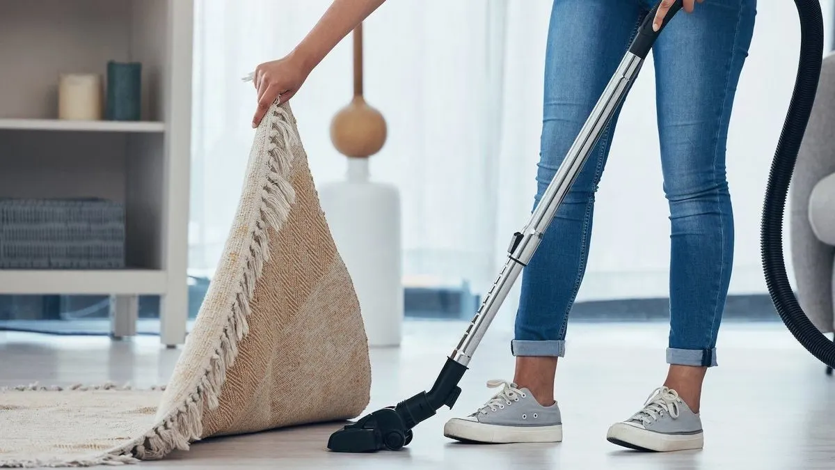 Person in Jeans und Turnschuhen saugt Holzboden, hebt einen beigen Teppich mit Fransen an; Regal und Sofa im Hintergrund.