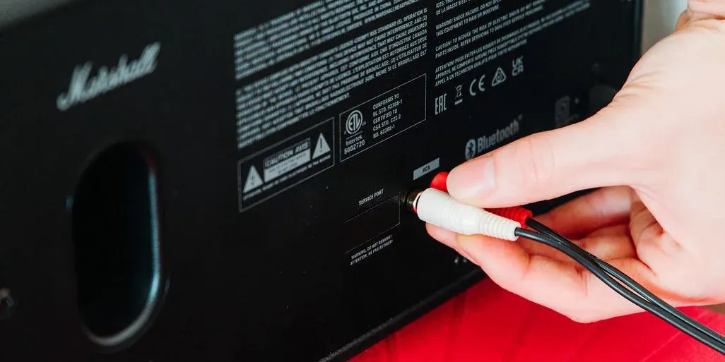 Eine Hand steckt ein weiß-rotes Cinch-Kabel in einen schwarzen Marshall-Lautsprecher. Bluetooth- und RCA-Anschlüsse sind sichtbar.