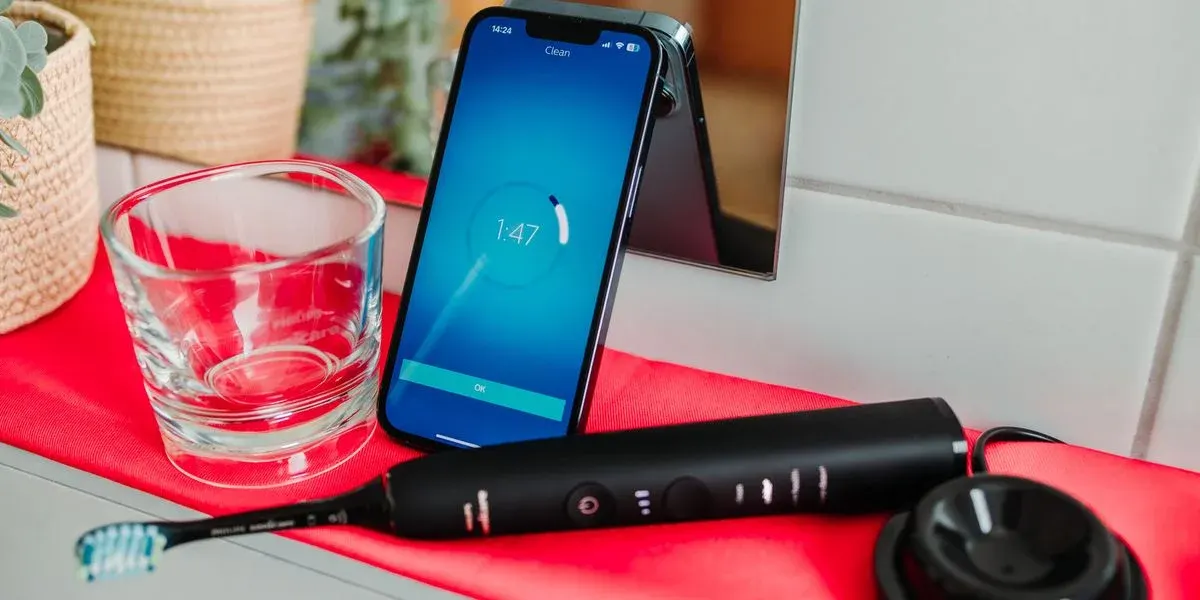 Schwarze elektrische Zahnbürste, klares Glas und Smartphone mit blauem Bildschirm (Timer 1:47) auf rotem Tuch. Weiße Fliesen, Korb im Hintergrund.