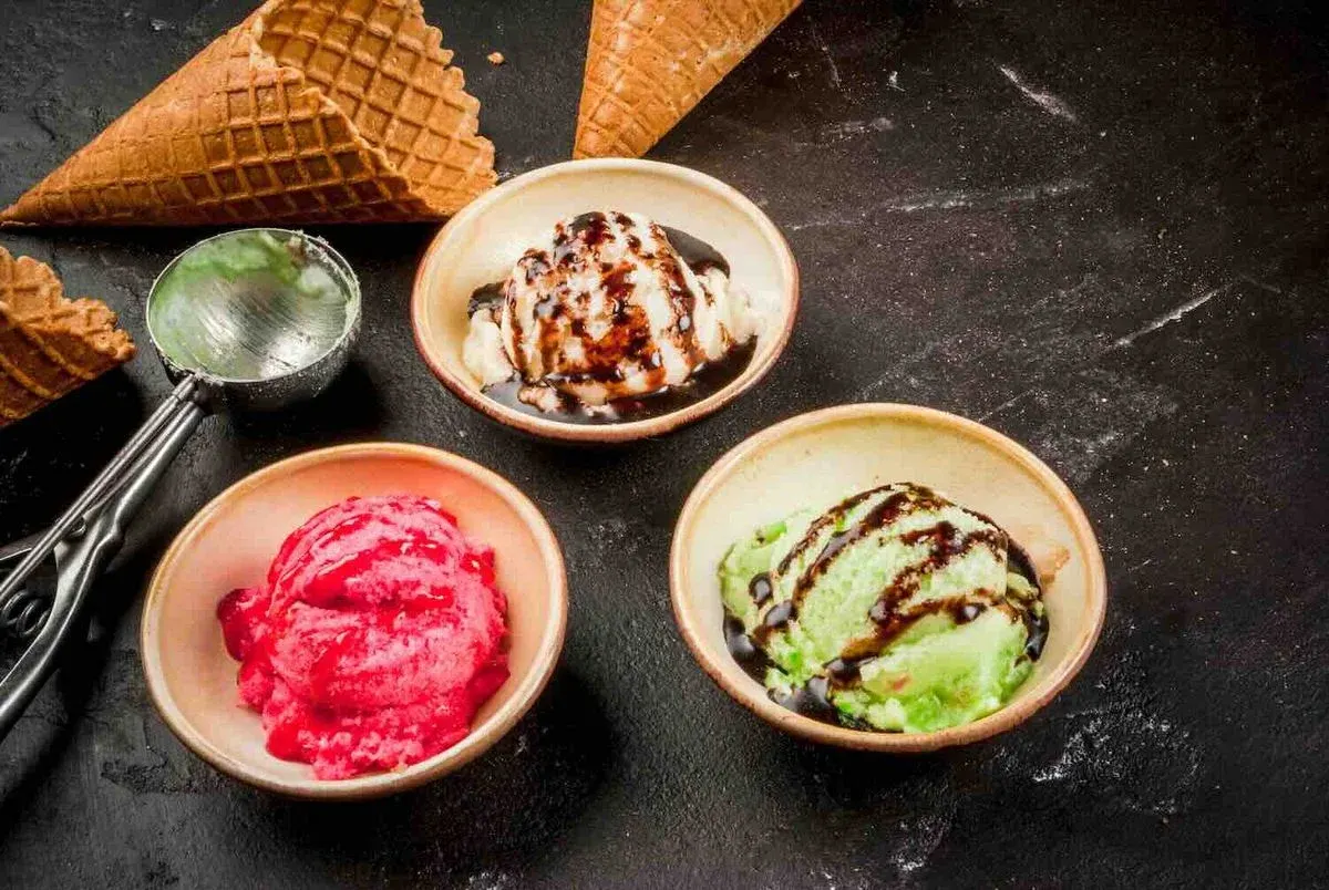 Drei bunte Eissorten in Schalen: Erdbeer-, Vanille- und Pistazieneis mit Schokoladensoße, zwei Waffelkegel und Portionierungsschaufel auf schwarzem Untergrund.