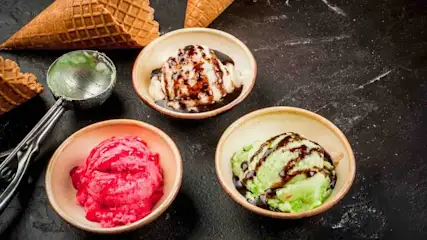 Drei bunte Eissorten in Schalen: Erdbeer-, Vanille- und Pistazieneis mit Schokoladensoße, zwei Waffelkegel und Portionierungsschaufel auf schwarzem Untergrund.