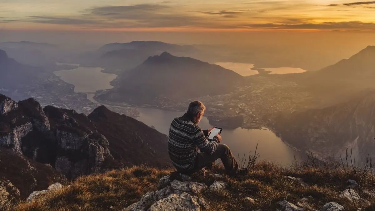 Mann sitzt auf einem Felsen am Berghang und liest auf einem e-Book Reader Im Hintergrund ist eine weite Aussicht auf einen See, Berge und eine Stadt bei Sonnenuntergang.