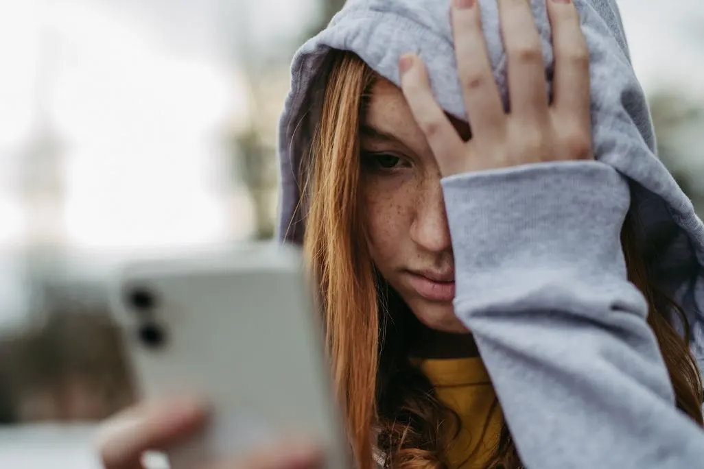 Eine Frau mit Kapuzenpullover hält ein Smartphone in der Hand und wirkt verzweifelt.