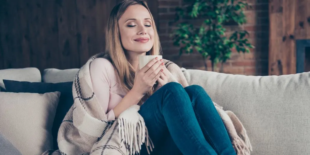 Eine Person sitzt entspannt auf einem hellgrauen Sofa, in eine beige Decke gehüllt, hält eine helle Tasse. Im Hintergrund sind eine Ziegelwand und eine Pflanze zu sehen.