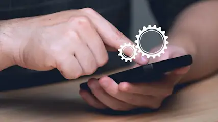 Eine Hand bedient ein Smartphone, über dem zwei Zahnräder als Symbol schweben.