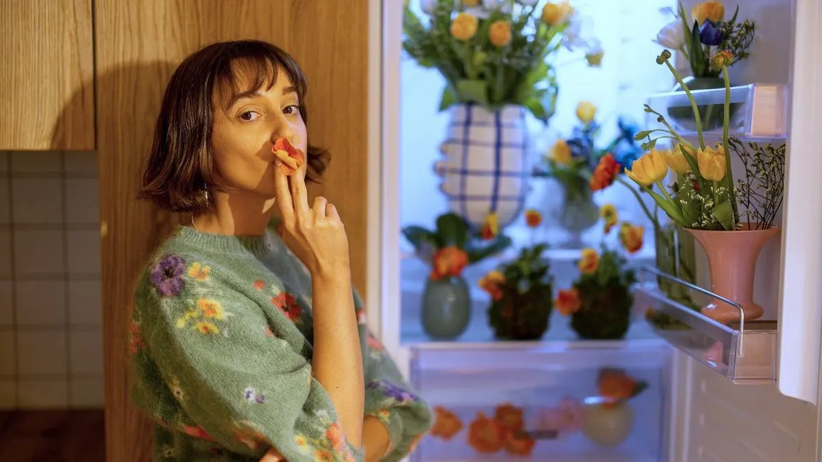 Eine Person in grünem Blumenpullover hält ein orangefarbenes Blütenblatt an die Lippen. Im Hintergrund ist ein Kühlschrank voller farbiger Blumen in Vasen.