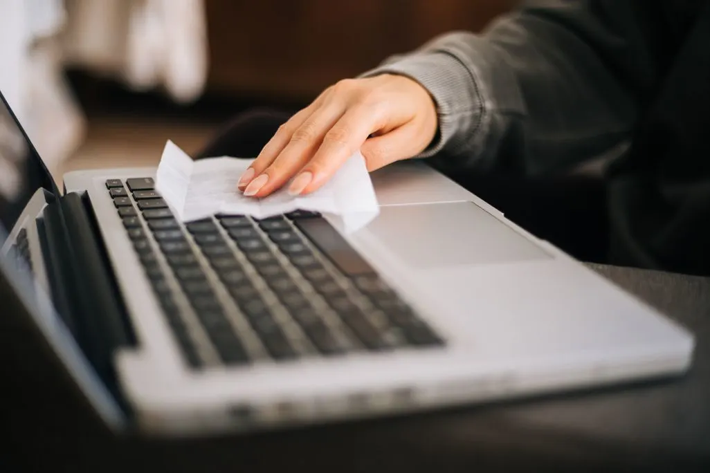 Hand wischt Laptop-Tastatur mit weißem Reinigungstuch sauber.