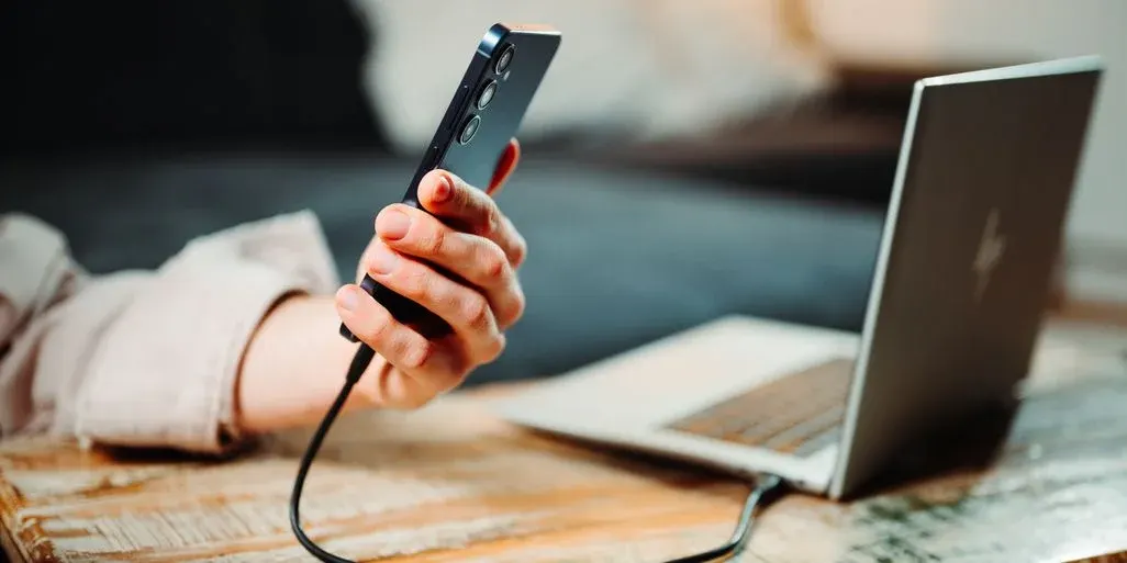 Hand hält Smartphone, dieses ist per Ladekabel mit Laptop verbunden, den man im Hintergrund sieht.