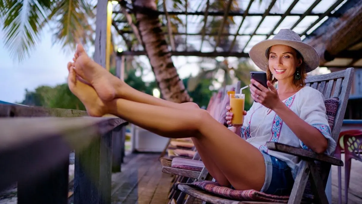 Frau sitzt auf einem Liegestuhl unter Palmen und benutzt ein Smartphone.