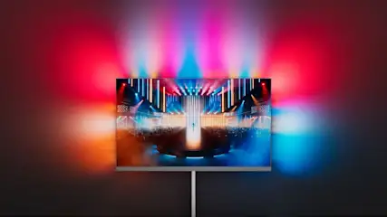 Philips OLED-Fernseher auf einem silbernen Standfuß, der eine Konzertszene mit bunter Bühnenbeleuchtung zeigt. Das Ambilight-System des Fernsehers projiziert die Bildschirmfarben – Rot, Pink, Blau und Orange – auf die dunkle Wand dahinter und erzeugt einen beeindruckenden Lichthalo-Effekt.