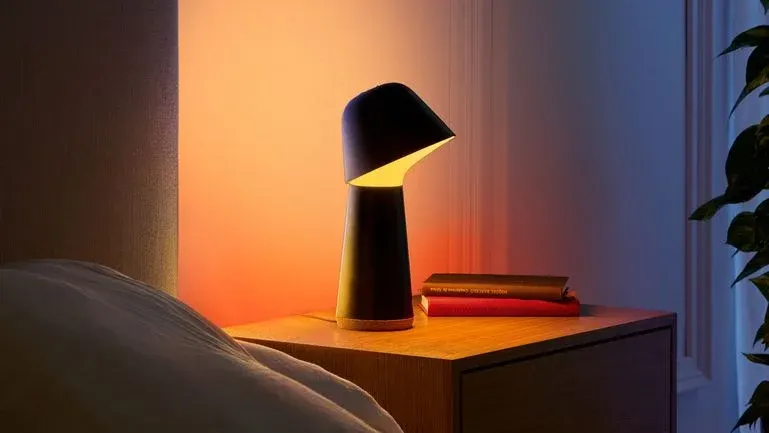 Eine dunkle Tischlampe mit warmem Licht steht auf einem Holznachttisch. Daneben liegen zwei Bücher. Links ist ein weißes Bett zu sehen.