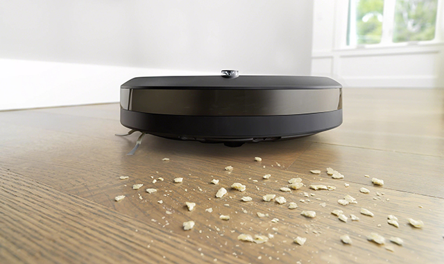 Ein schwarzer Staubsaugerroboter reinigt einen Holzboden mit hellen Krümeln. Im Hintergrund sind weiße Wände und ein Fenster sichtbar.