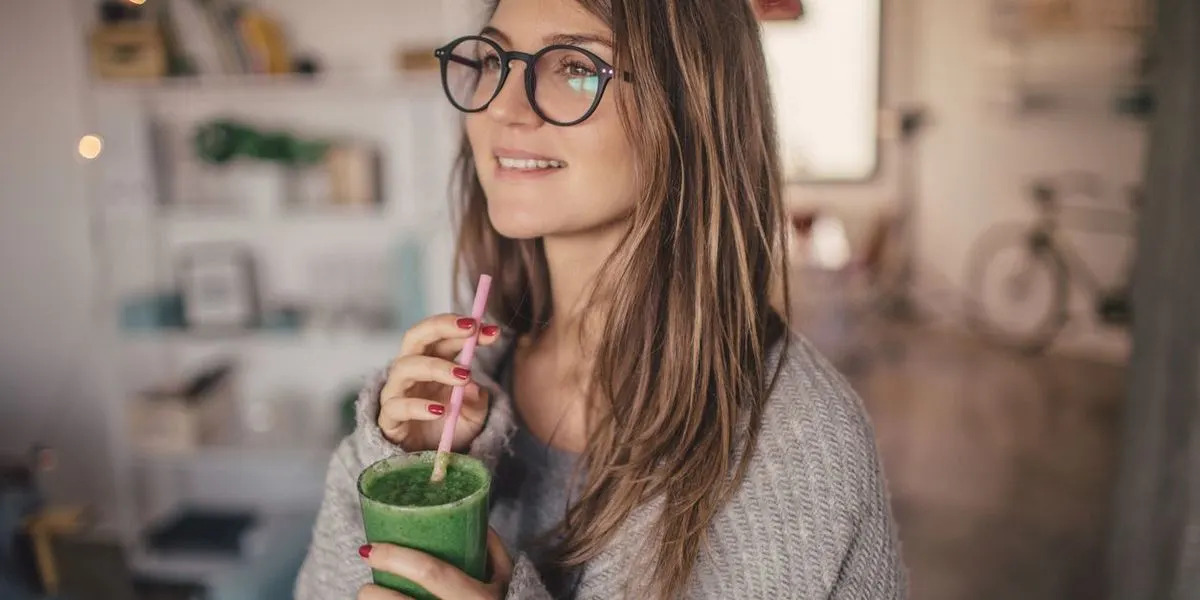 Frau mit langen Haaren und Brille trägt einen grauen Strickpullover und hält einen grünen Smoothie im Glas mit Strohhalm in der Hand.