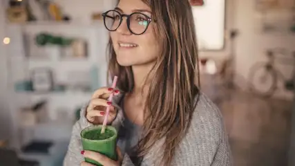 Frau mit langen Haaren und Brille trägt einen grauen Strickpullover und hält einen grünen Smoothie im Glas mit Strohhalm in der Hand.