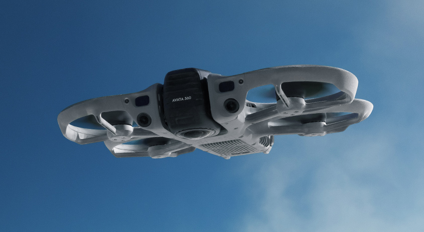 Nahaufnahme der DJI Avata 360 Drohne im Flug von unten vor blauem Himmel, Fokus auf Kamera und Rotoren für 360°-Aufnahmen.