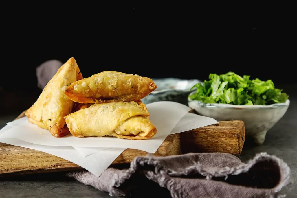 Drei knusprige Samosas auf Backpapier angerichtet mit Kräutern im Hintergrund.
