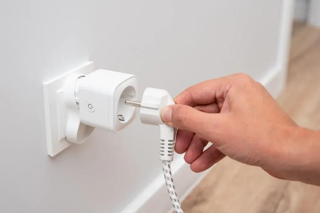 Eine männliche Hand hält einen weißen Schukostecker mit einem schwarz-weiß geflochtenen Kabel und steckt ihn in einen smarten Zwischenstecker (Smart Plug), der bereits in einer wandmontierten Steckdose sitzt. Der Smart Plug ist weiß und verfügt über einen sichtbaren Power-Button mit einem Ein-/Aus-Symbol. Die Wand ist weiß, im Hintergrund ist ein heller Holzboden zu erkennen.