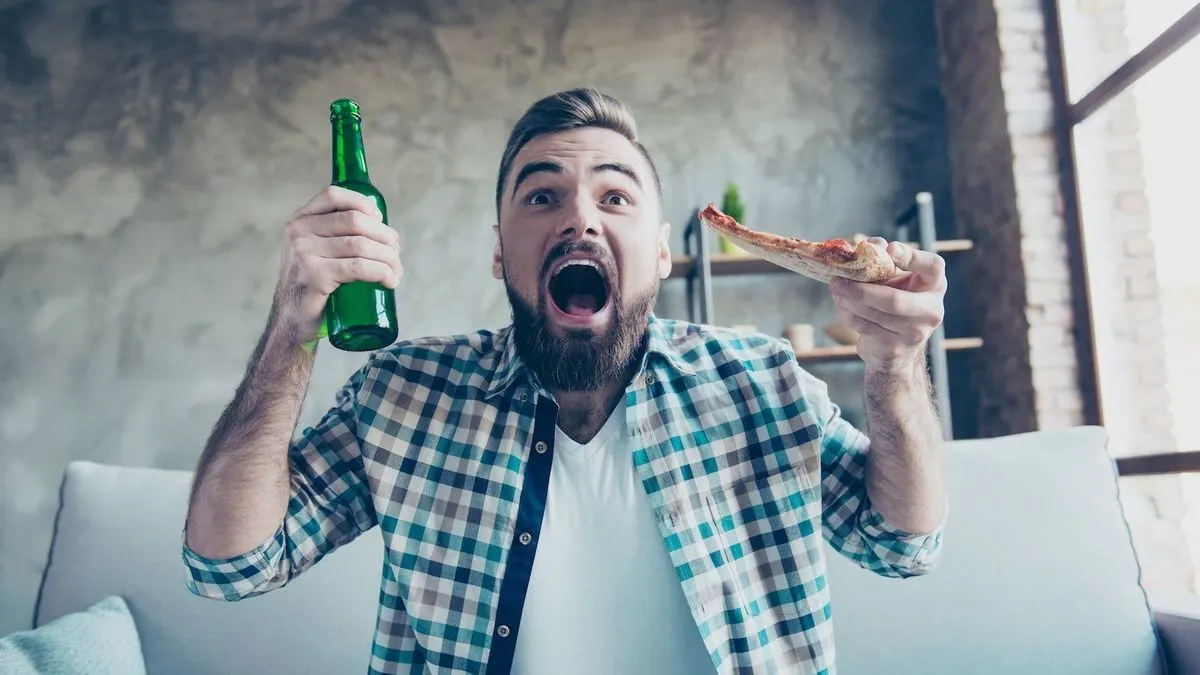 Ein aufgeregter Mann im karierten Hemd hält eine Flasche und ein Stück Pizza in den Händen.
