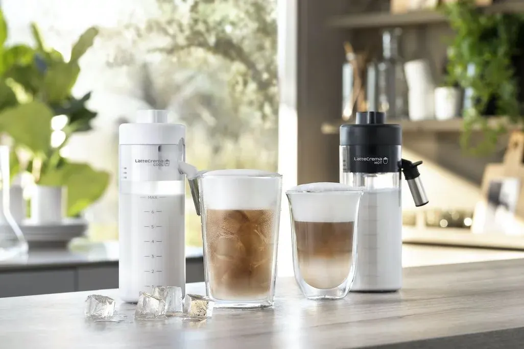 Zwei Milchaufschäumer, weiß und schwarz, stehen mit zwei Kaffeegetränken, eines mit Eis und Schaum, auf Holztheke. Eiswürfel im Vordergrund. Hintergrund: Küche mit Pflanzen.