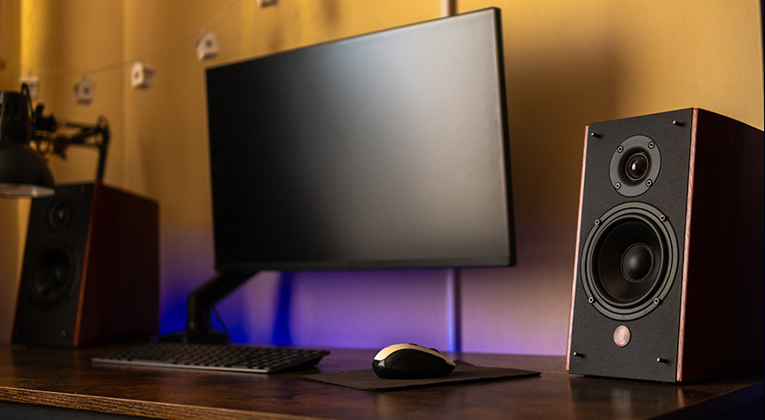 Ein Schreibtisch mit einem Monitor, einer Tastatur, einer Maus und zwei Lautsprechern vor einer gelben Wand.