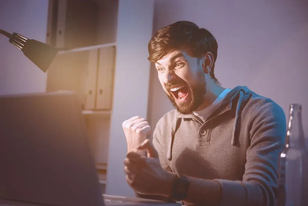 Junger Mann sitzt vor einem Laptop und jubelt mit geballter Faust; sein Gesicht zeigt überschwängliche Freude, warmes Lampenlicht fällt auf ihn.
