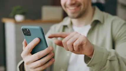 Eine Person in einem grünen Hemd hält ein blaugrünes Smartphone mit drei Kameralinsen und tippt auf den Bildschirm.