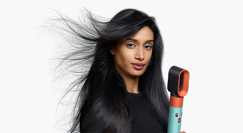 Eine Person mit wehendem, langem, schwarzem Haar hält einen Haarstyler vor einem weißen Hintergrund.