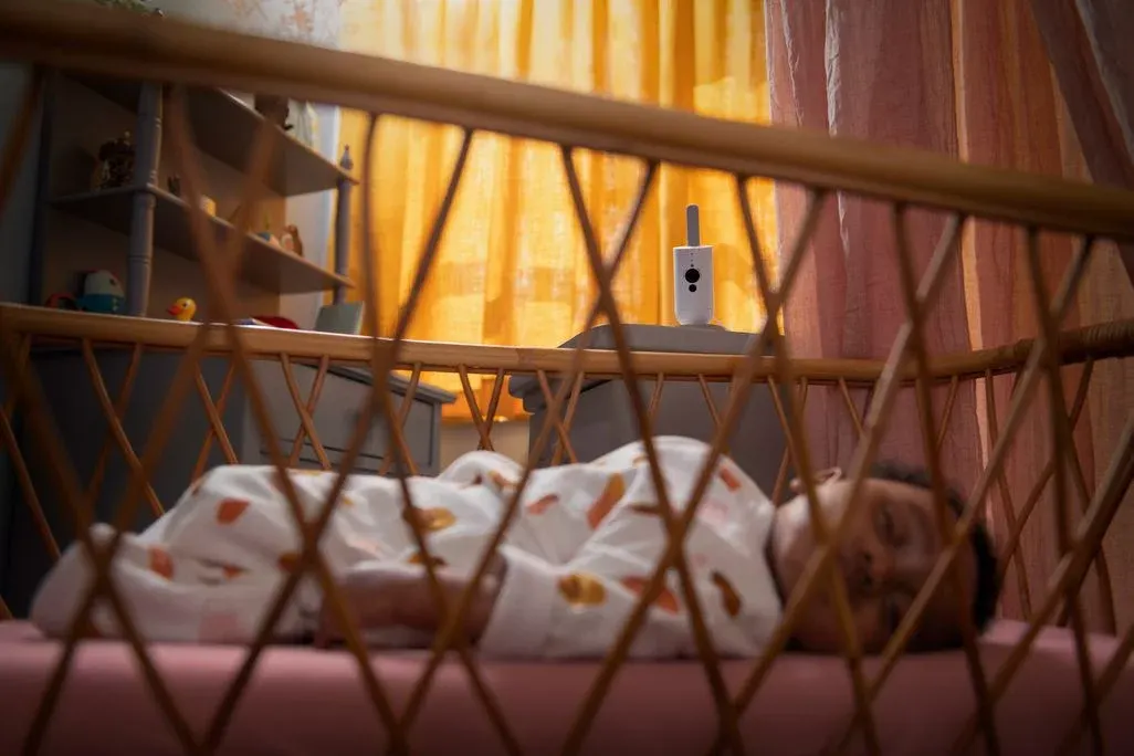 Schlafendes Baby im Gitterbett, im Hintergrund steht auf einer Kommode eine weiße Babycam vor gelbem Vorhang.