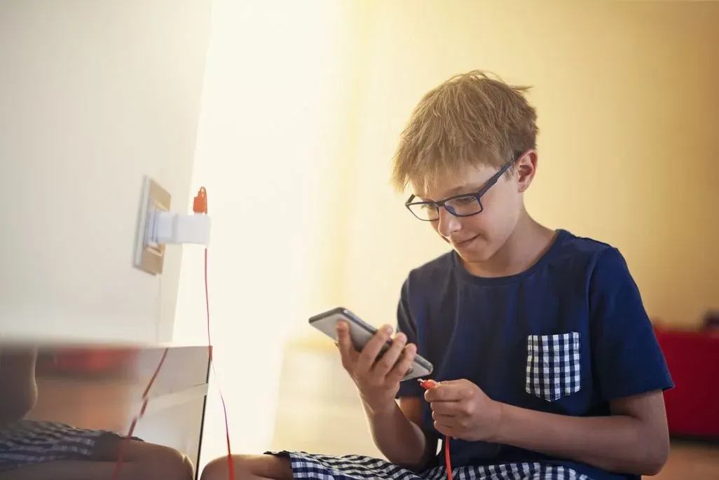 Junge mit Brille sitzt auf dem Boden und schaut auf sein Smartphone, daneben eine Steckdose mit rotem Ladekabel.