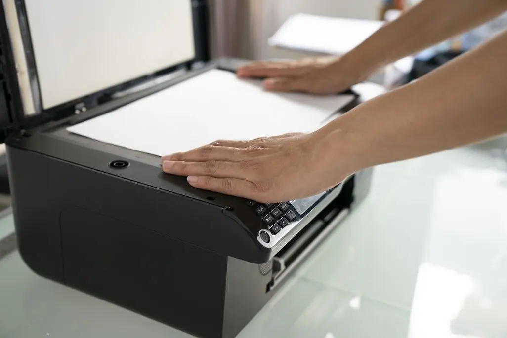 Zwei Hände legen weißes Papier auf das geöffnete Scan-Bett eines schwarzen Druckers/Scanners auf einem hellen Tisch.