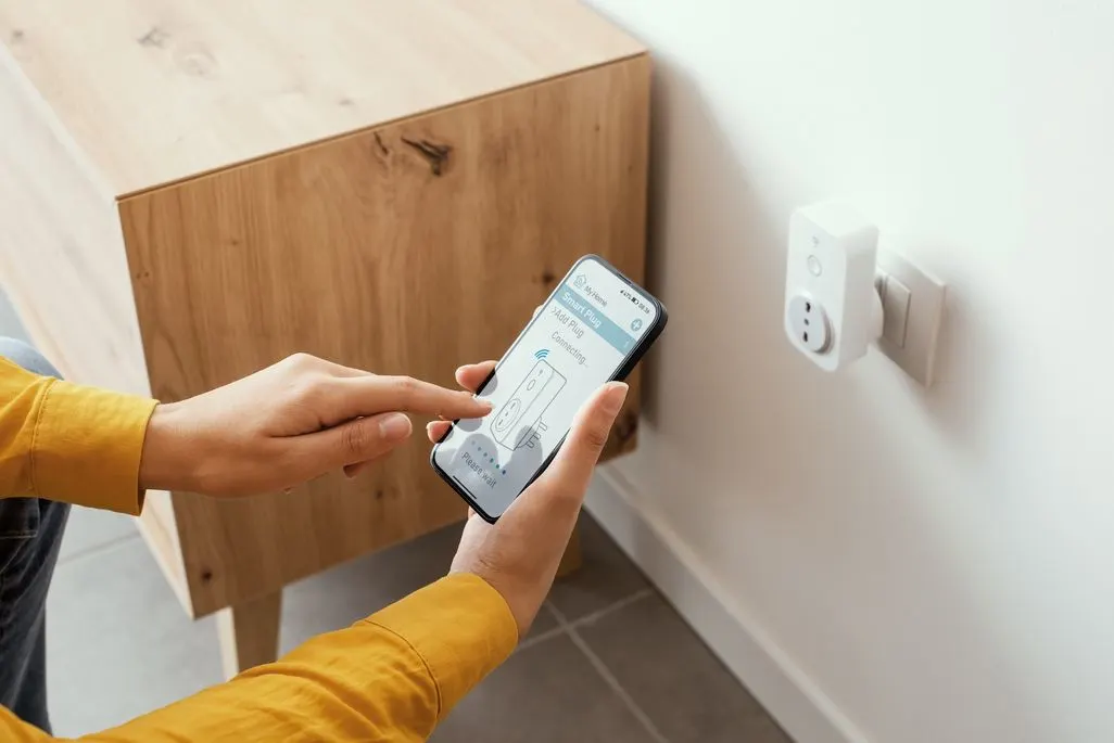 Person bedient eine Smart Home App auf dem Smartphone vor einer intelligenten Steckdose an der Wand.