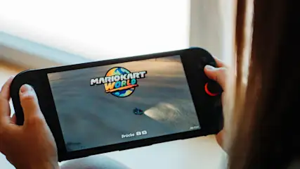 Person hält schwarze Nintendo Switch. Bildschirm zeigt "Mario Kart World" Logo, blauen Kart auf grauer Straße, "Drücke L R".