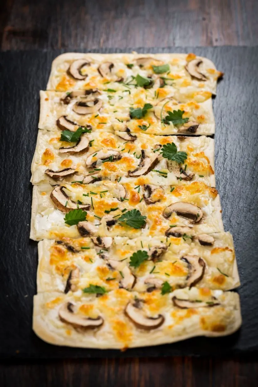 Flammkuchen mit Pilzen und Kräutern auf dunklem Untergrund.