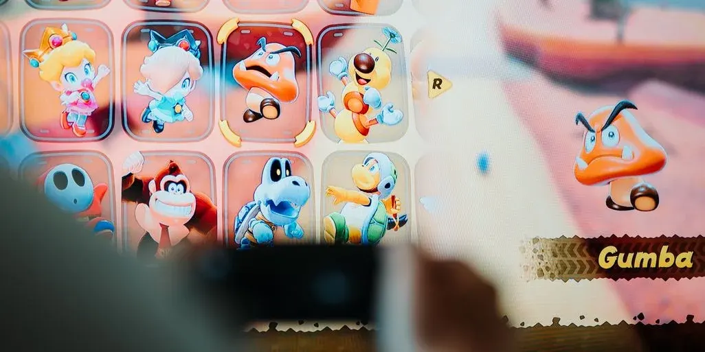 Videospiel-Charakterauswahl mit bunten Figuren wie Baby Peach und Donkey Kong. Ein großer, wütender orangefarbener Goomba ist ausgewählt. Hände halten Controller.