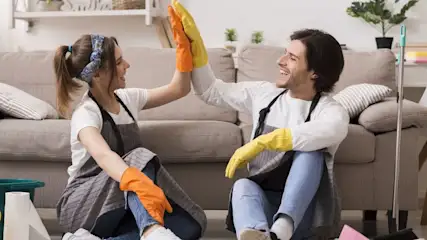 Zwei Personen sitzen in einem Wohnzimmer und geben sich ein High Five beim gemeinsamen Putzen.