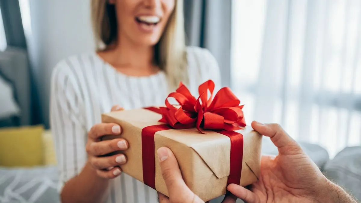 Einer lächelnden Frau wird ein Geschenk mit roter Schleife überreicht.