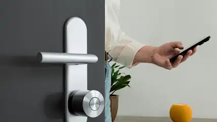Eine Person mit einem Smartphone in der Hand geht durch eine geöffnete dunkle Tür, auf der das Nuki Smart Lock Ultra montiert ist.