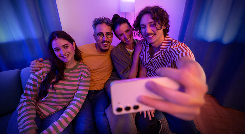 Junge Menschen sitzen in einer Gruppe zusammen und lächeln in die Kamera des Smartphones.